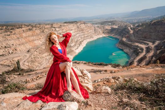 A beautiful girl in a red long dress, Sits on a rock high above the lake in the afternoon. Against the background of the blue sky and the lake in the shape of a heart