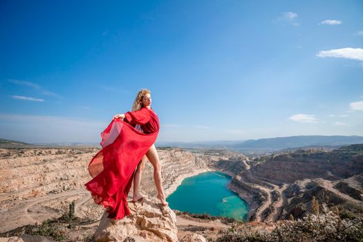 Side view of a beautiful sensual woman in a red long dress posing on a rock high above the lake in the afternoon. Against the background of the blue sky and the lake in the form of a heart.