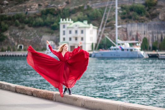 A woman in a red dress, a fashion model with long silk wings in a flowing dress, flying fabric on the embankment