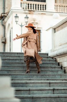 Outdoor fashion portrait of young elegant fashionable brunette woman, model in stylish hat, choker and light raincoat posing at sunset in European city