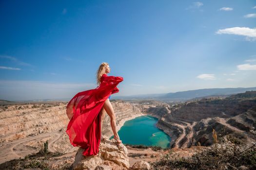 Side view of a beautiful sensual woman in a red long dress posing on a rock high above the lake in the afternoon. Against the background of the blue sky and the lake in the form of a heart.