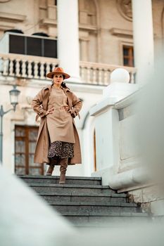 Outdoor fashion portrait of young elegant fashionable brunette woman, model in stylish hat, choker and light raincoat posing at sunset in European city