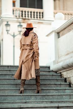 Outdoor fashion portrait of young elegant fashionable brunette woman, model in stylish hat, choker and light raincoat posing at sunset in European city