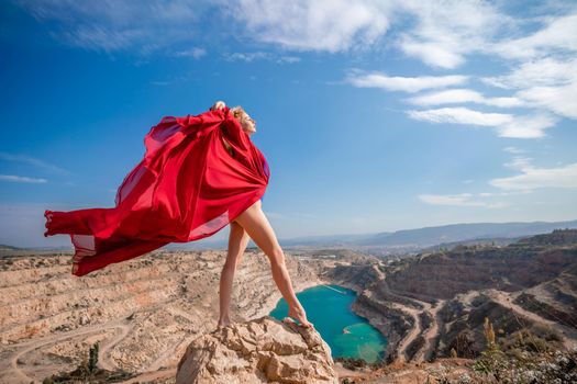 Side view of a beautiful sensual woman in a red long dress posing on a rock high above the lake in the afternoon. Against the background of the blue sky and the lake in the form of a heart.