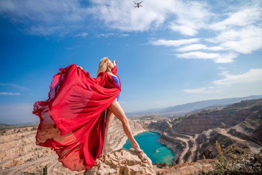 Side view of a beautiful sensual woman in a red long dress posing on a rock high above the lake in the afternoon. Against the background of the blue sky and the lake in the form of a heart.