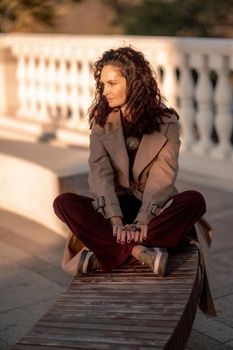 Outdoor fashion portrait of young elegant fashionable brunette woman, model in stylish hat, choker and light raincoat posing at sunset in European city