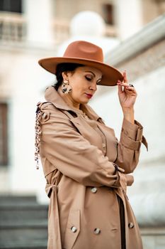 Outdoor fashion portrait of young elegant fashionable brunette woman, model in stylish hat, choker and light raincoat posing at sunset in European city