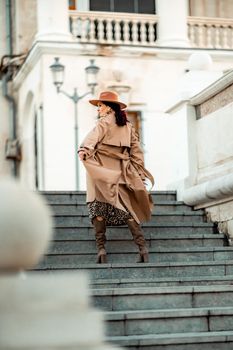 Outdoor fashion portrait of young elegant fashionable brunette woman, model in stylish hat, choker and light raincoat posing at sunset in European city