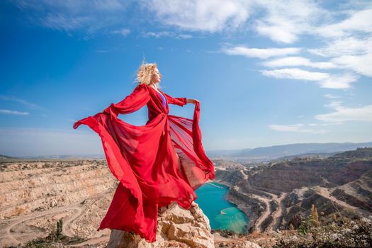 Side view of a beautiful sensual woman in a red long dress posing on a rock high above the lake in the afternoon. Against the background of the blue sky and the lake in the form of a heart.