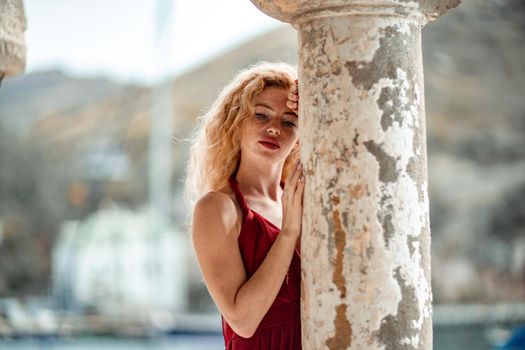 Outdoor portrait of a young beautiful natural redhead girl with freckles, long curly hair, in a red dress, posing against the background of the sea