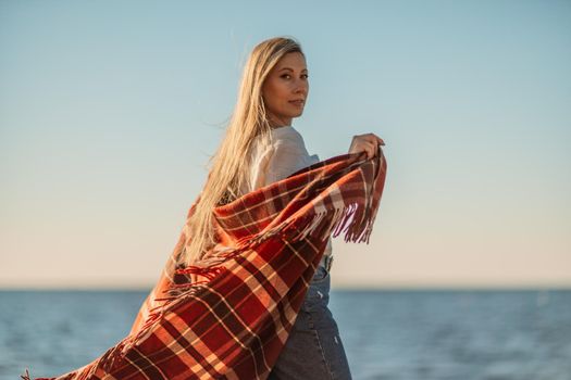 Attractive blonde Caucasian woman enjoying time on the beach at sunset, walking in a blanket and looking to the side, with the sunset sky and sea in the background. Beach vacation.