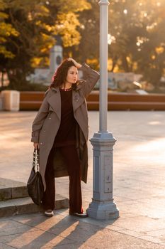 Outdoor fashion portrait of young elegant fashionable brunette woman, model in stylish hat, choker and light raincoat posing at sunset in European city