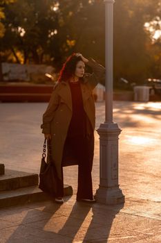 Outdoor fashion portrait of young elegant fashionable brunette woman, model in stylish hat, choker and light raincoat posing at sunset in European city