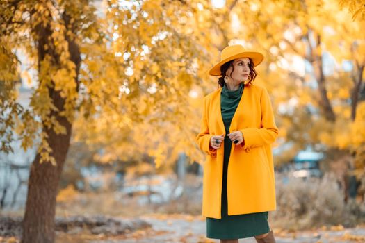 Beautiful woman walks outdoors in autumn. She is wearing a yellow coat, yellow hat and green dress. Young woman enjoying the autumn weather. Autumn content.