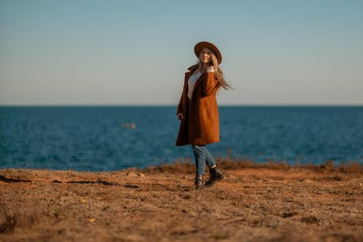 A woman walking along the coast near the sea. An elegant lady in a brown coat and a hat with fashionable makeup walks on the seashore.