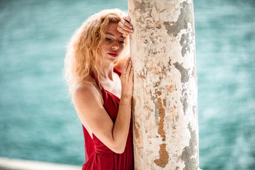 Outdoor portrait of a young beautiful natural redhead girl with freckles, long curly hair, in a red dress, posing against the background of the sea