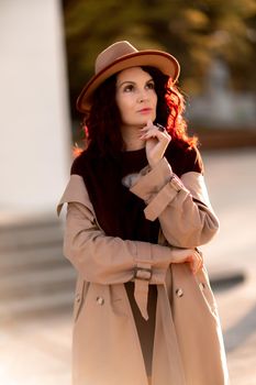 Outdoor fashion portrait of young elegant fashionable brunette woman, model in stylish hat, choker and light raincoat posing at sunset in European city