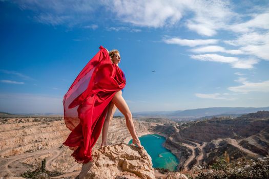 Side view of a beautiful sensual woman in a red long dress posing on a rock high above the lake in the afternoon. Against the background of the blue sky and the lake in the form of a heart.