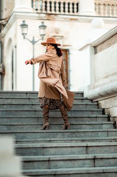 Outdoor fashion portrait of young elegant fashionable brunette woman, model in stylish hat, choker and light raincoat posing at sunset in European city