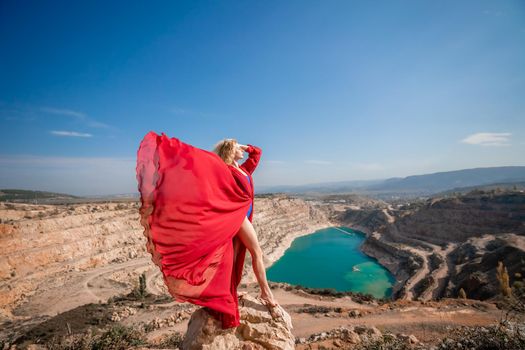 Side view of a beautiful sensual woman in a red long dress posing on a rock high above the lake in the afternoon. Against the background of the blue sky and the lake in the form of a heart.