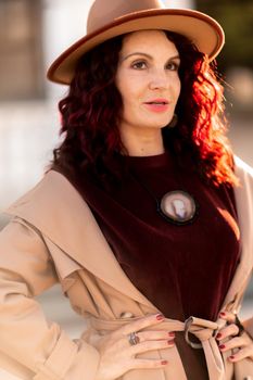 Outdoor fashion portrait of young elegant fashionable brunette woman, model in stylish hat, choker and light raincoat posing at sunset in European city