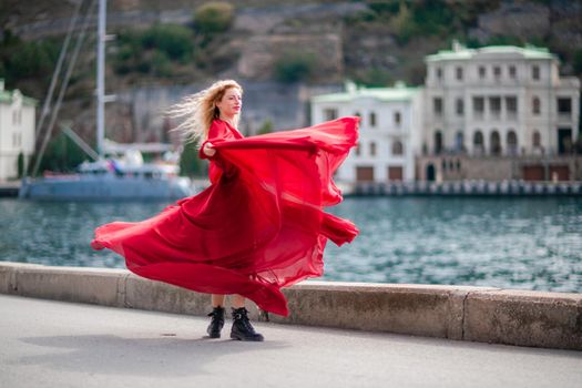 A woman in a red dress, a fashion model with long silk wings in a flowing dress, flying fabric on the embankment