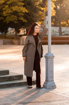 Outdoor fashion portrait of young elegant fashionable brunette woman, model in stylish hat, choker and light raincoat posing at sunset in European city