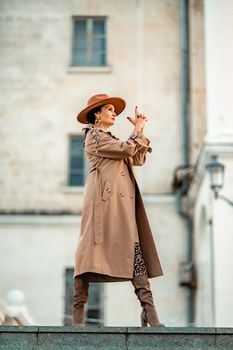 Outdoor fashion portrait of young elegant fashionable brunette woman, model in stylish hat, choker and light raincoat posing at sunset in European city