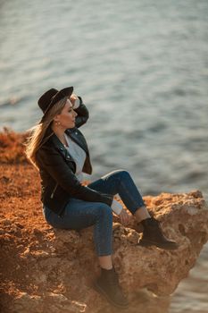 A serious blonde girl in a stylish black leather jacket and a black hat is sitting with her back to the seashore.
