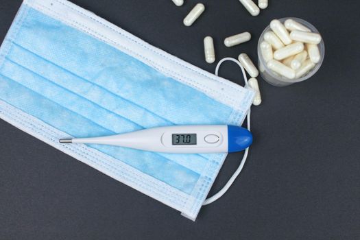 White electronic thermometer with packs of tablets and blue medical face mask on black background.