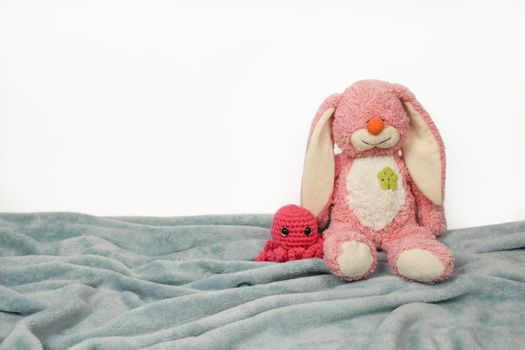 A plush bunny toy sitting on plush blanket and white wall. Plush pink bunny toy with pink knitted octopus.