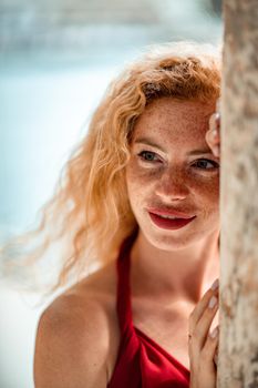 Outdoor portrait of a young beautiful natural redhead girl with freckles, long curly hair, in a red dress, posing against the background of the sea