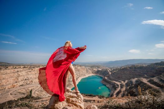 Side view of a beautiful sensual woman in a red long dress posing on a rock high above the lake in the afternoon. Against the background of the blue sky and the lake in the form of a heart.