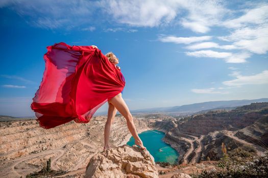 Side view of a beautiful sensual woman in a red long dress posing on a rock high above the lake in the afternoon. Against the background of the blue sky and the lake in the form of a heart.