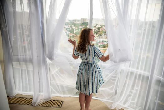 Rear view of a young joyful woman wearing fashion dress and holding the curtains open to look out of large light window at home, turning to look and smile at camera, interior.
