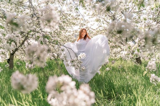 Fantasy woman in long white elegant fashion long dress walks in green spring blossom cherry garden. Happy cheerful girl princess bride. Skirt fabric flies flowing waving in wind motion