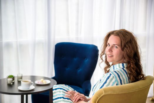 Young woman at home sitting on modern chair in front of window relaxing in her living room and drinking coffee or tea with sweets or desert. Relax before new business day.