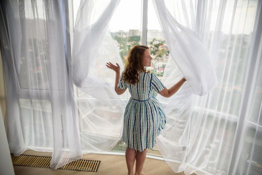 Rear view of a young joyful woman wearing fashion dress and holding the curtains open to look out of large light window at home, turning to look and smile at camera, interior.