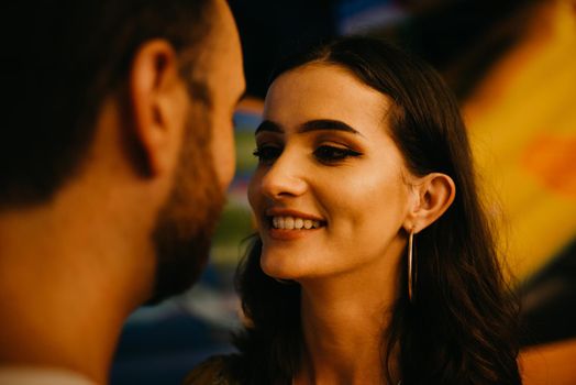 A close portrait of a woman in a yellow dress with a plunging neckline who is admiring her boyfriend with a beard near amusement rides. A couple of lovers on a date at the fair in Valencia.