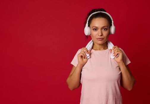 Confident portrait of an attractive young Hispanic fit sporting woman with wireless headphones and white terry towel in pink t-shirt looking at camera isolated over red background with copy ad space