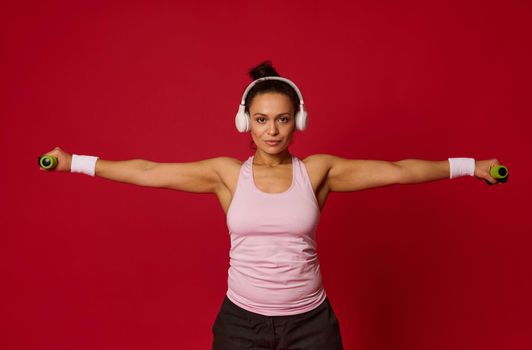 Studio shot of a beautiful muscular multiethnic woman wearing a sports top and white wireless headphones on a red background listening to music and exercising with dumbbells