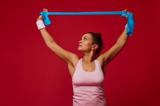 Pleasant charming African American active woman doing bodyweight training with resistance elastic rubber band on red background with copy space for ads. Fitness, workout, stretching exercises.