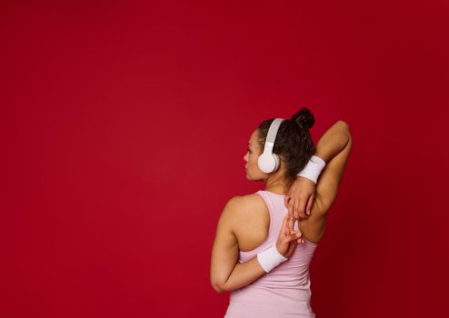 Rear view of a determined multi-ethnic female athlete with headphones stretching arms while exercising against red colored background with advertising copy space