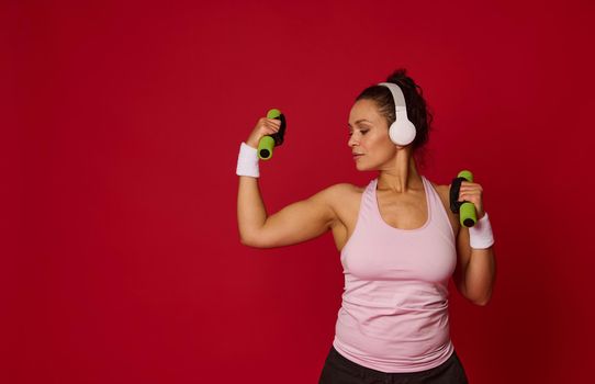 Attractive determined confident young Hispanic athletic woman in headphones, tight pink sportswear and white terry bracelets doing arm exercises with dumbbells on red background with copy ad space