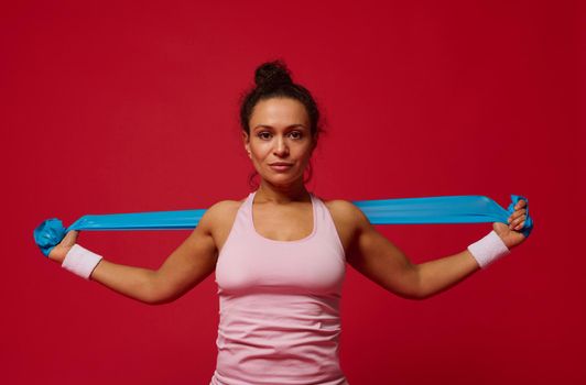 Beautiful confident motivated sportswoman working out with blue elastic band on red background and looking at camera. Copy ad space