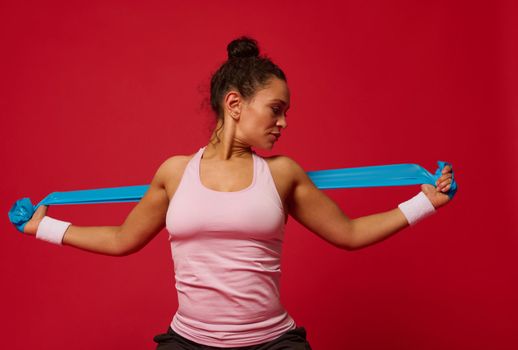 African American determined middle-aged muscular woman in tight pink top and terry wristbands doing stretching exercises on arms using elastic fitness band against red background with copy ad space