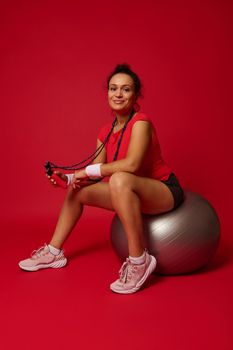 Beautiful Hispanic athletic woman in red tight top and black sports shorts holding skipping rope and smiling at camera while sitting on silver fit ball on red background with copy ad space