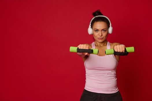 Determined athletic woman with white wireless headphones doing exercises on her arms, holding small dumbbells at outstretched hands isolated on a red background with space for advertising text