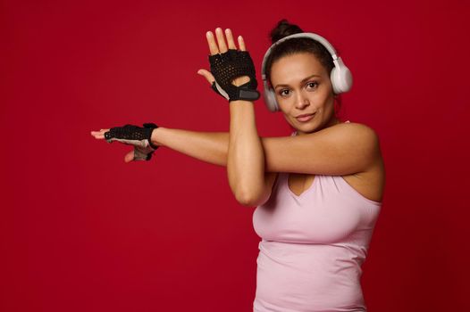 Confident active middle-aged active multi-ethnic woman in sportswear and headphones stretching arms, looking at camera against red background with copy ad space