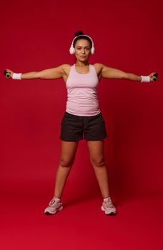 Full length body size view of female athlete exercising with dumbbells against red background with copy space for advertising text. Sport, fitness, healthy and active lifestyle, weight loss concept
